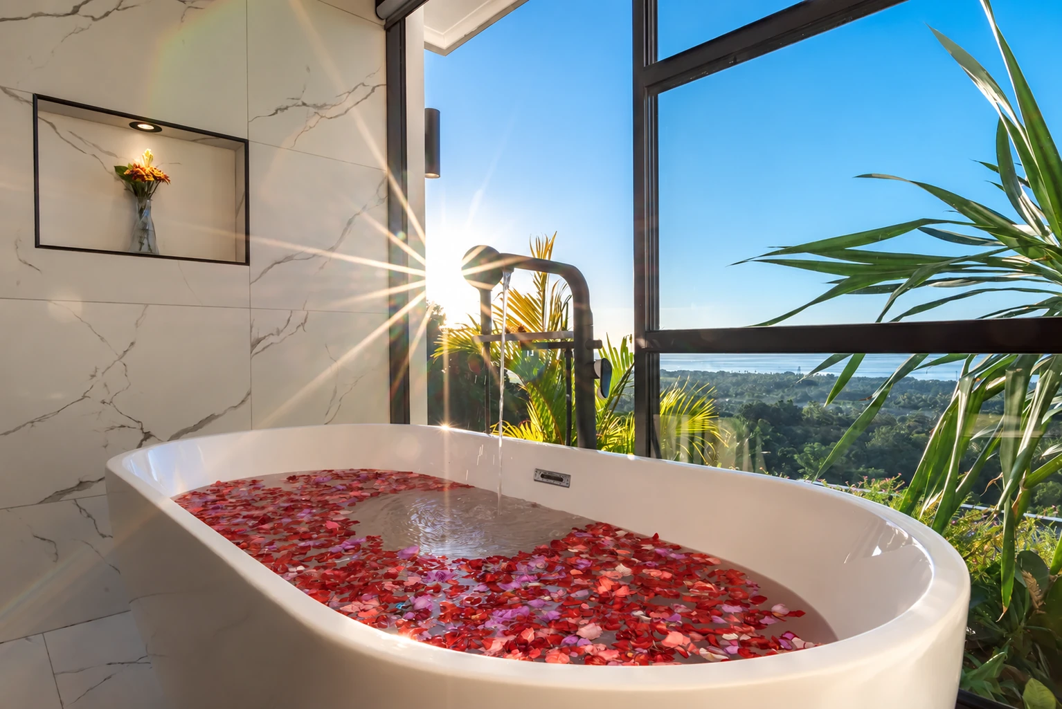 Freestanding bath with flower petals and sea-view window in warm sunlight.