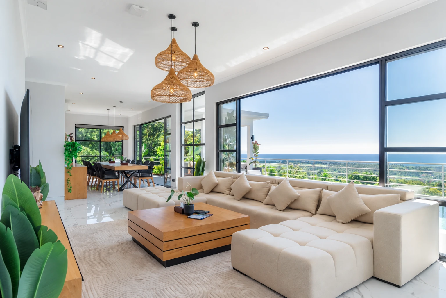 Open living room with cream sectional sofa, timber table, pendant lights and broad sea-facing windows.