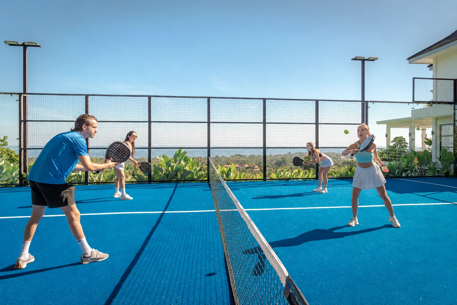 Four adults playing doubles on blue padel court at the property, sea visible behind.